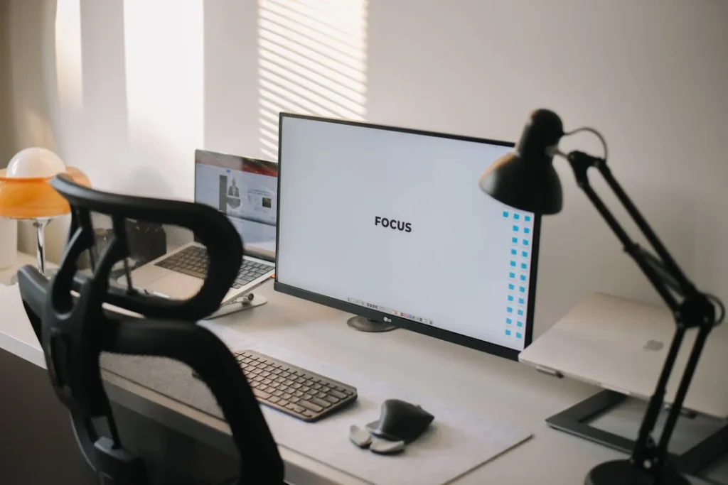 A monitor on a desk with a chair and a lamp.